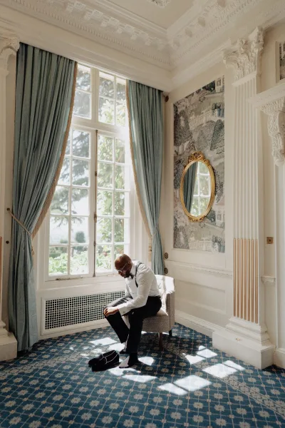 Groom getting ready, putting on loafers by a sunlit window with ornate molding, a wallpaper mural, and patterned carpet.