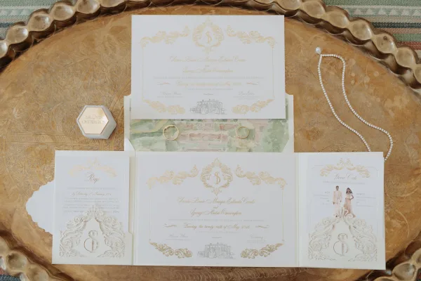 Wedding invitation suite with gold foil invitations arranged on a brass tray over a woven textile, accented by paired wedding bands.
