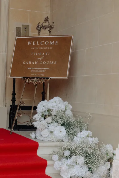 Wedding welcome sign on an ornate easel set beside stone steps and a stone wall, greeting arriving guests.