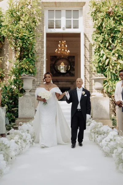 Ceremony processional with the bride walking down aisle, holding a white rose bouquet toward a grand mansion entrance with a chandelier beyond.