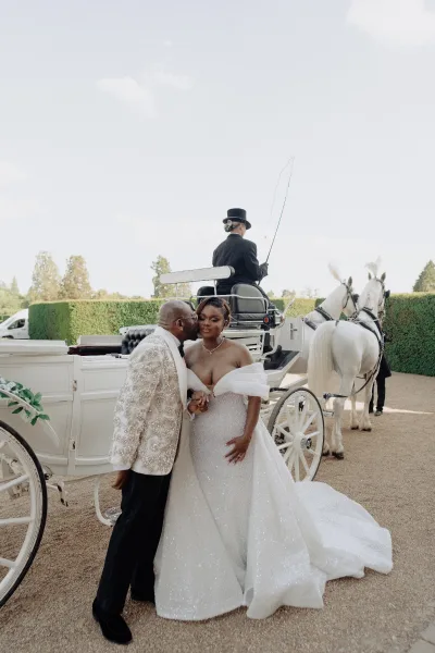 Couple portrait of a bride and groom with a horse-drawn carriage, sharing a cheek kiss and clasped hands in formal wedding attire.