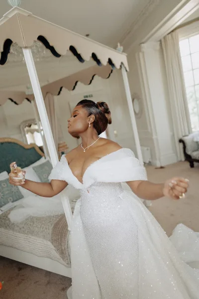 Bride getting ready, bride spraying perfume from a glass bottle by a canopy bed, soft window light and curtains set a calm bedroom scene.