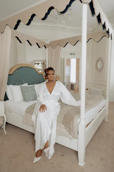 Bridal portrait of a bride on bed in a satin bridal robe, seated on a canopy bed with a tufted headboard and soft curtains in the background.