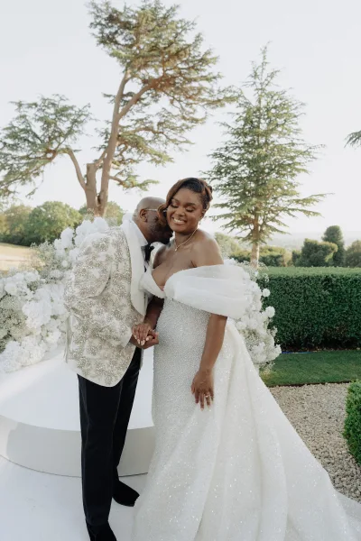 Couple portrait with the groom kissing the bride's cheek, his embellished tuxedo jacket catching light, along a garden path framed by hedges.