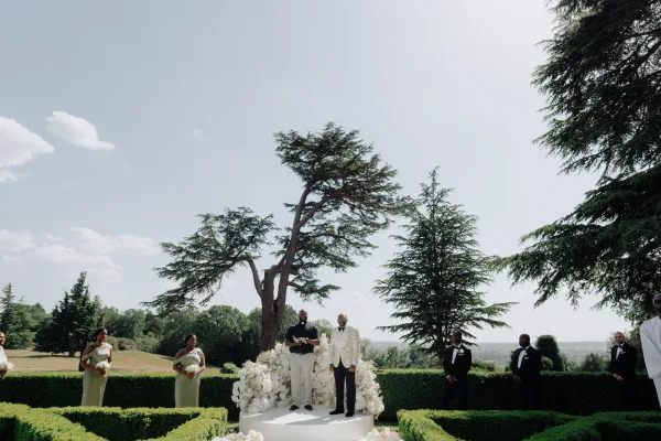 Ceremony moment at an outdoor wedding ceremony as the groom stands at the altar, framed by a white floral backdrop beneath tall garden trees.