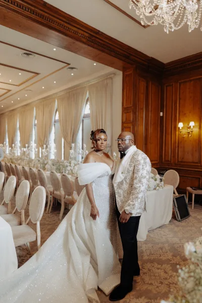 Couple portrait, a ballroom wedding portrait with bride in an off-the-shoulder ball gown beside groom with glasses beneath a crystal chandelier.
