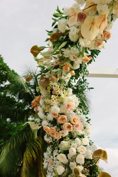 Wedding floral arch with asymmetrical wedding arch flowers in peach and white, featuring roses and anthurium, set against sky and palm trees