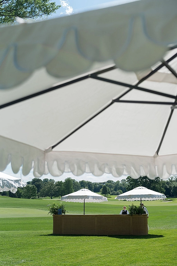 Outdoor bar setup with a bar counter and white patio umbrellas, greenery arrangements, and a small sign on a lawn under blue sky