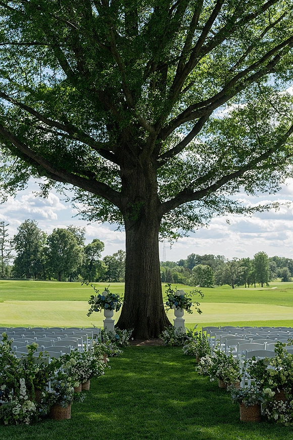 Outdoor ceremony setup with white folding chairs lining a floral wedding aisle, arranged beneath a large tree on a grassy lawn