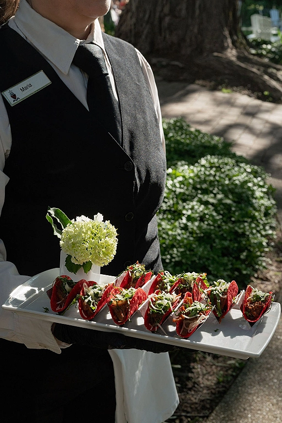 Wedding cocktail appetizers on a white tray, passed hors d'oeuvres wedding by a server in black vest and tie in a sunny garden walkway