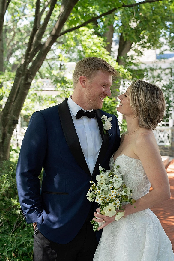 Couple portrait of bride and groom smiling, her strapless lace dress and white bouquet beside his navy tuxedo on a brick garden path