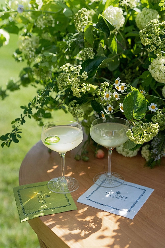 Wedding cocktail detail with signature wedding cocktails in coupe glasses, citrus garnish and greenery on a wooden side table on a garden lawn