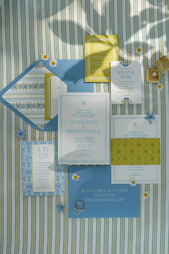 Invitation suite wedding invitation flat lay with envelopes, RSVP and rehearsal cards, rings in a box, and daisies on striped fabric