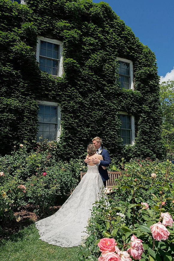 Wedding kiss portrait of bride and groom kissing beside a wooden bench, her lace gown train flowing against an ivy-covered building backdrop
