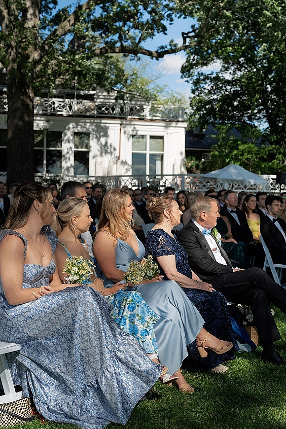 Wedding guests seated in folding chairs during an outdoor ceremony, with blue bridesmaid dresses and bouquets on a sunny lawn by trees