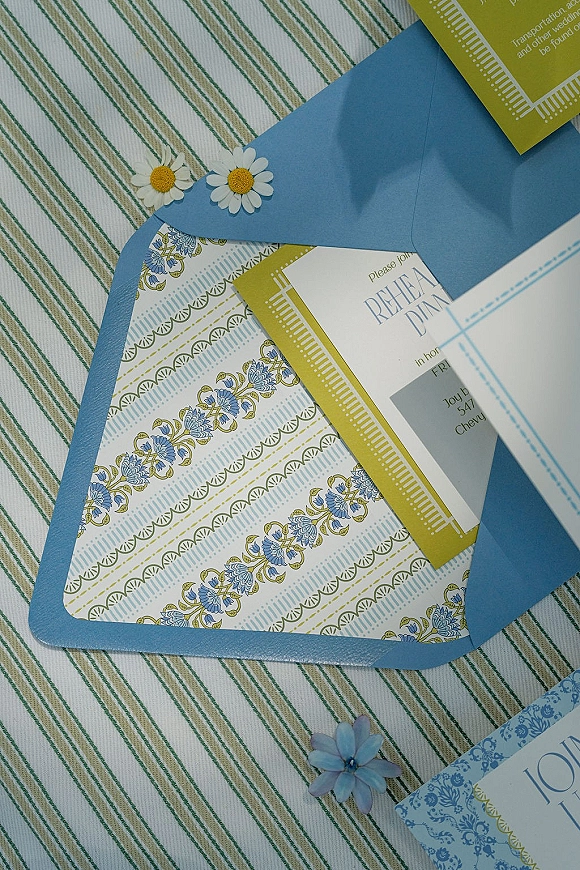 Wedding invitations with a blue wedding invitation suite, patterned card, RSVP, and daisy embellishments on a striped fabric surface