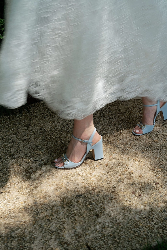 Bridal shoes in light blue wedding shoes with ankle straps and metallic buckles, peeking from lace dress hem on a sunlit gravel path