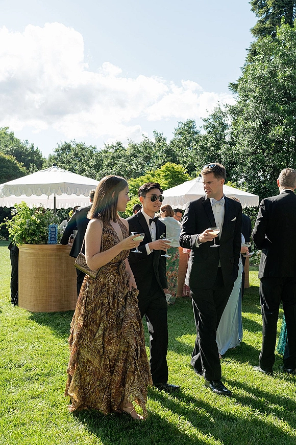 Wedding cocktail hour with guests mingling in tuxedos and satin dress, sipping coupe cocktails at an outdoor bar on a sunny lawn under umbrellas