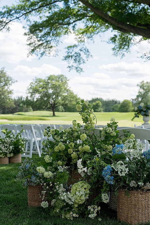 Outdoor ceremony decor with white folding chairs lining a grassy aisle, hydrangea and greenery arrangements in woven baskets under trees