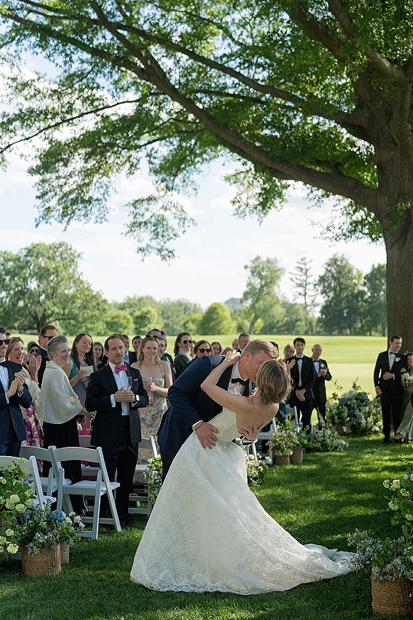 Wedding kiss as the bride dips back in her dress and groom in a tuxedo holds her, guests lining a chair-filled aisle beneath a tree canopy