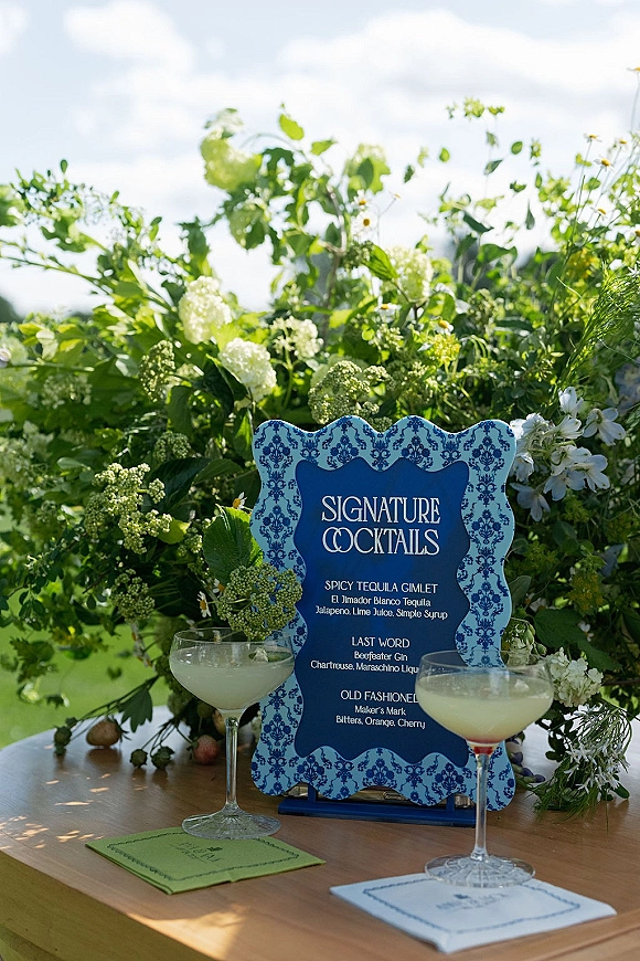 Signature cocktail sign listing wedding signature cocktails on a wooden table with green-and-white florals, coupe glasses, and garden lawn behind