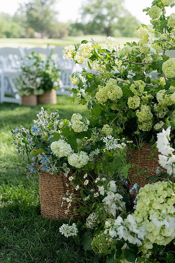 Ceremony aisle flowers in woven baskets with white hydrangeas, wildflowers, and greenery lining an outdoor lawn aisle by chairs and trees