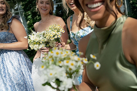 Bridesmaids portrait with bridesmaids holding bouquets of white and yellow daisies, in mix-and-match dresses beside a sunlit greenhouse