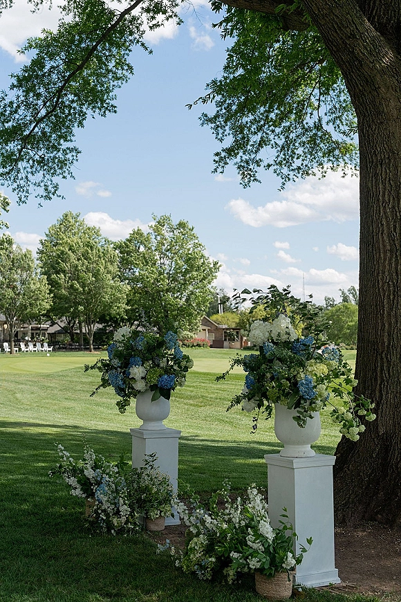 Ceremony floral arrangements frame the entrance on white pedestal stands, with blue and white urn blooms beneath a large tree on a lawn