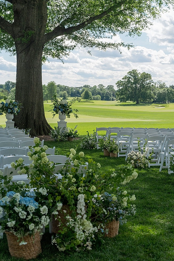 Outdoor ceremony setup with wedding ceremony chairs lining a flower-lined aisle of hydrangeas and greenery under a large tree on a lawn