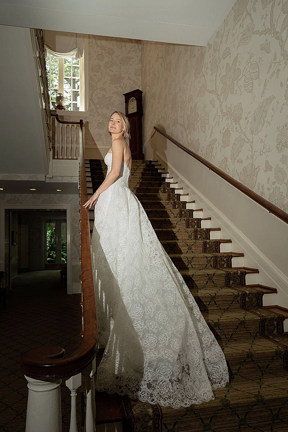 Bridal portrait of a bride on staircase in a strapless lace wedding dress with long train, holding the banister in a floral hallway
