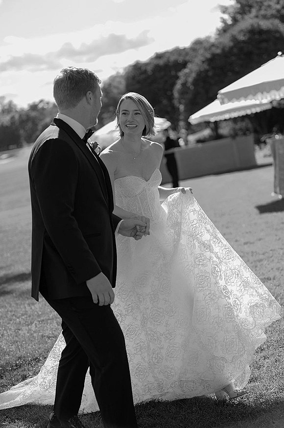 Wedding couple portrait in black and white as the bride and groom hold hands walking on a lawn, bride lifting her train near an outdoor tent