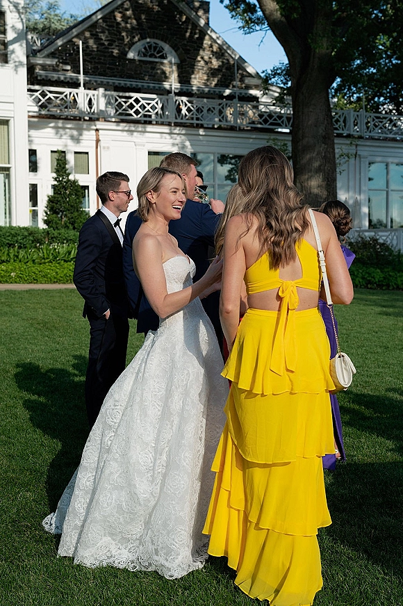 Wedding cocktail hour as the bride mingles with guests on a green lawn, lace ballgown among tuxedos and a yellow dress by a white estate