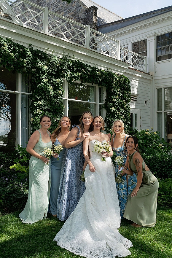 Bridesmaid group photo of bride with bridesmaids holding bouquets, with lace gown and ivy-covered white house wall backdrop outdoors