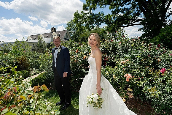 Couple portrait in a rose garden, bride in a strapless gown holding a white bouquet, groom in tuxedo smiling under cloudy sky