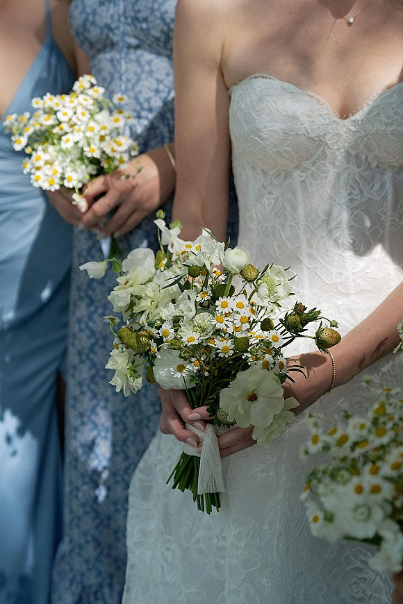 Bridal bouquet of chamomile daisies and white flowers with greenery, held against a lace sweetheart dress in warm outdoor sunlight