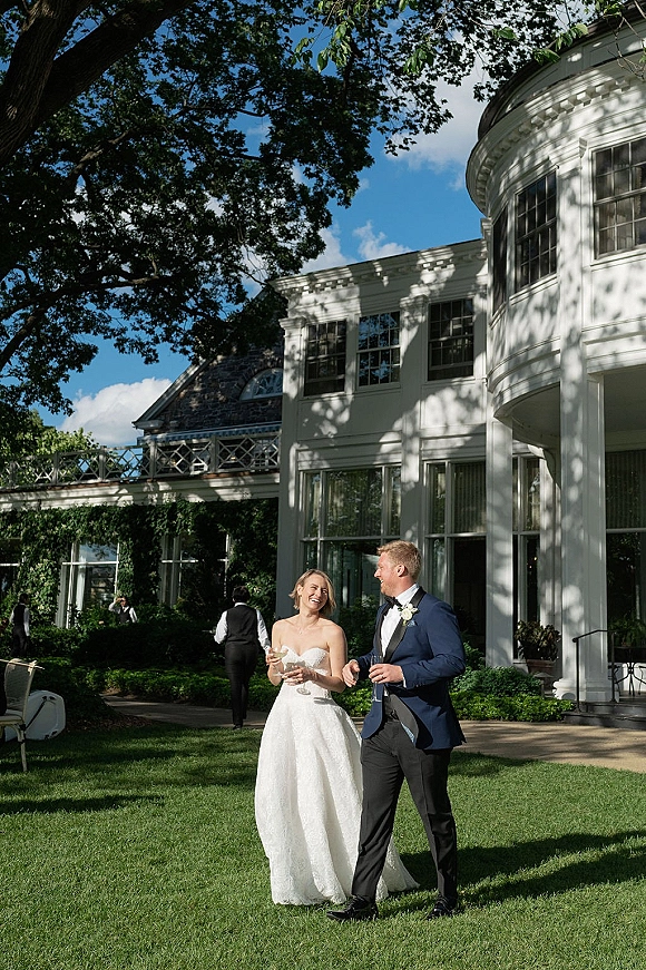 Couple portrait of bride and groom laughing, walking with wine glasses on a garden lawn beside a large white house under blue sky