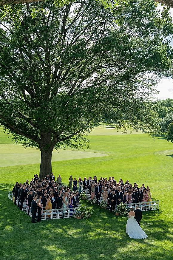 Outdoor wedding ceremony beneath a large tree, with white folding chairs in a circle as the bride walks the flower-lined aisle on a lawn