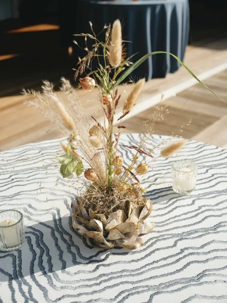 Wedding centerpiece with dried flower arrangement and pampas grass, votive candles on a striped tablecloth against a sunlit dark curtain backdrop