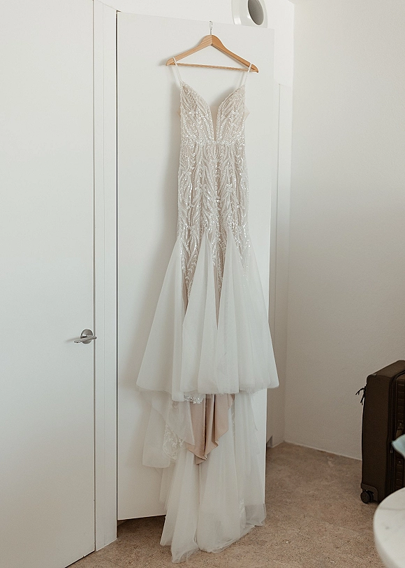 Wedding dress with a beaded wedding dress bodice and plunge neckline hanging on a wooden hanger against a white door and wall