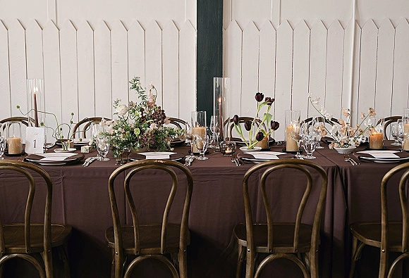 Reception tablescape with a long wedding head table set in brown linen, white and green florals, tall cylinder and votive candles against paneled walls