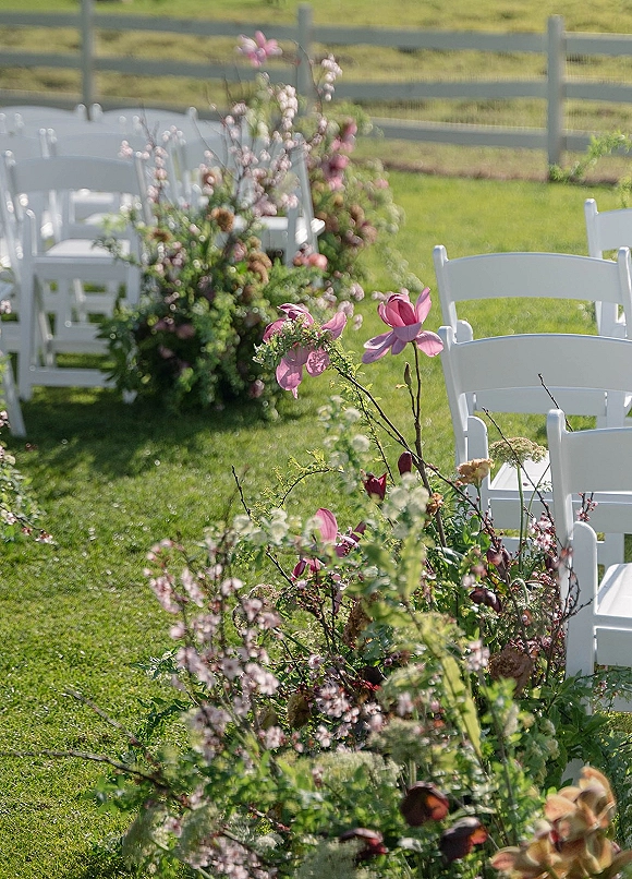Ceremony aisle decor with outdoor ceremony aisle lined by pink flowers and greenery beside white folding chairs on a grass lawn near a wooden fence