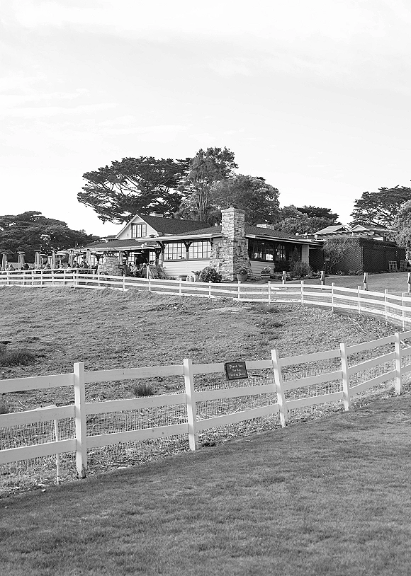 Wedding venue exterior with a white fence, patio umbrellas, and outdoor tables near a stone chimney, set beside a grassy field and trees
