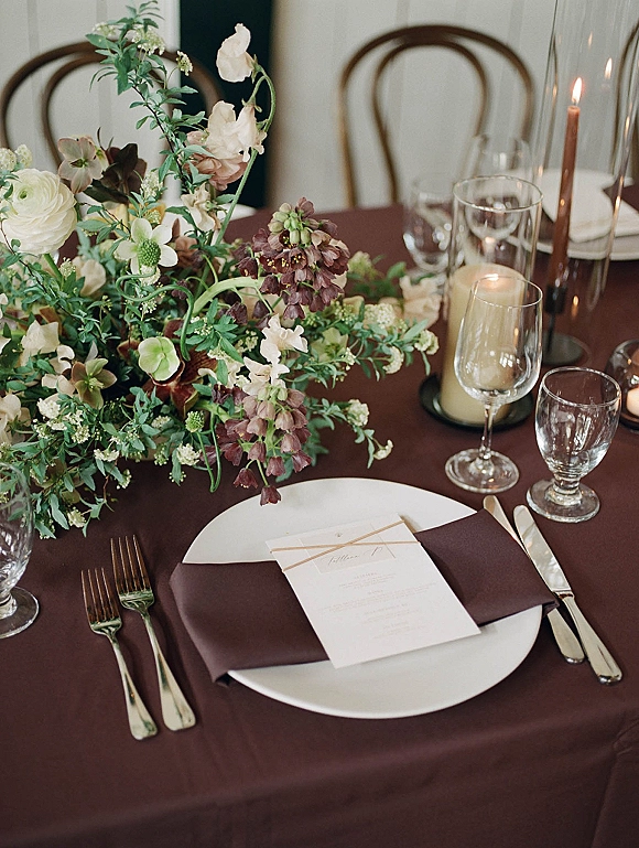 Reception tablescape wedding table setting with floral centerpiece, greenery, taper and pillar candles, plates, napkins, and glassware indoors