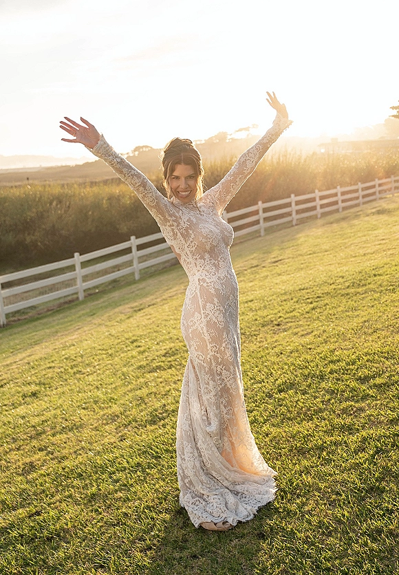 Bridal portrait of a bride smiling with arms raised, wearing a long sleeve lace wedding dress in a sunset field by a white fence