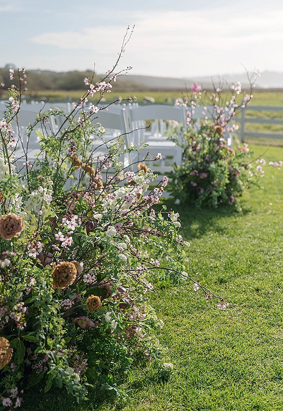 Ceremony aisle flowers with flowering branch accents and lush greenery beside white chairs on a grass lawn, with fence and distant hills beyond