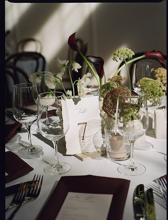 Reception tablescape with wedding table number card, calla lilies and sweet peas centerpiece, burgundy napkins, glassware, and votive candle in window light