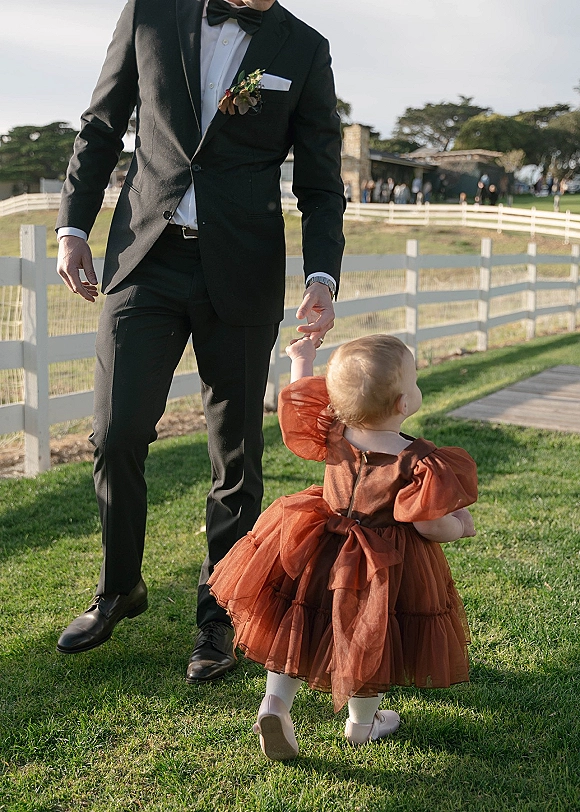 Wedding family moment as groom in black tuxedo walks hand in hand with a flower girl in a rust dress on a grassy lawn by a white fence