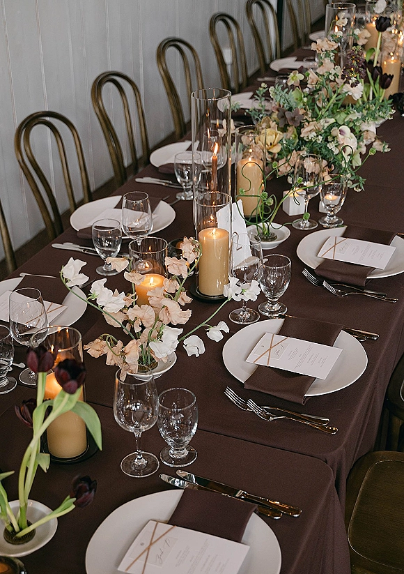 Reception tablescape with brown wedding table linen, white plates, gold flatware, menus, glassware, and candlelit tulip floral centerpieces on a long banquet table
