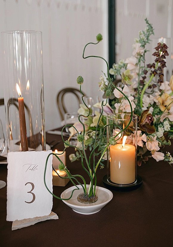 Reception tablescape with wedding table centerpiece, greenery and blush blooms, pillar and taper candles in glass hurricanes on a dark wood table