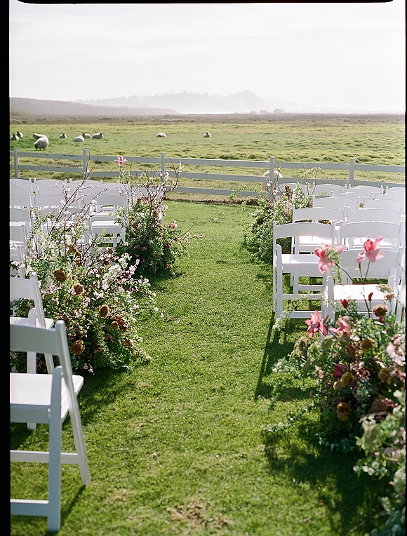 Outdoor ceremony aisle with wedding aisle flowers lining a grassy lawn, flanked by white folding chairs and a ranch fence with sheep beyond